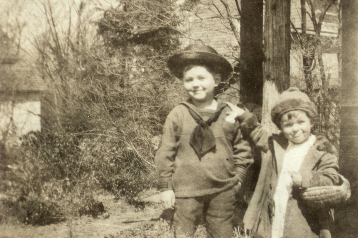 Original black and white photograph. Shows Teddy Ryder standing with one hand in his pocket. Beside him stands his sister, Margaret, with her hand on his shoulder.